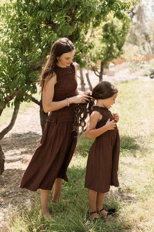 A woman braids a young girl’s hair outside on a sunny day. Both wear matching Ivy City Co Simone Dresses in Warm Cocoa, featuring a comfortable smocked bodice that adds style and maternity-friendly ease as they stand on green grass near leafy trees.