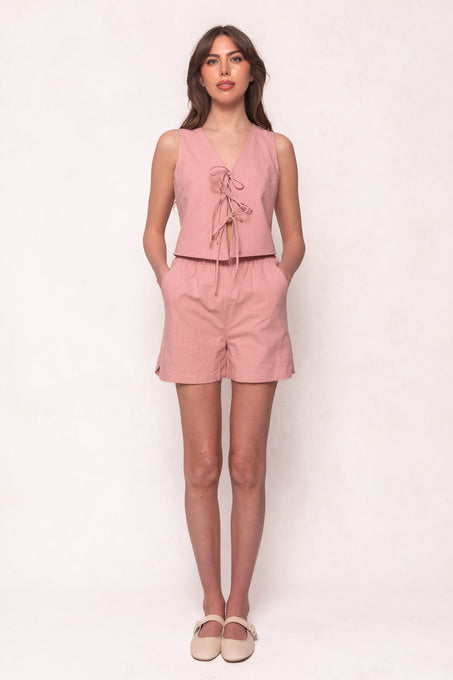 A woman with wavy brown hair wears Ivy City Cos Skye Shorts in Pink—high-waisted linen-cotton shorts with an elastic waistband—paired with a matching sleeveless pale pink v-neck front-tie top and beige flats, against a light background.