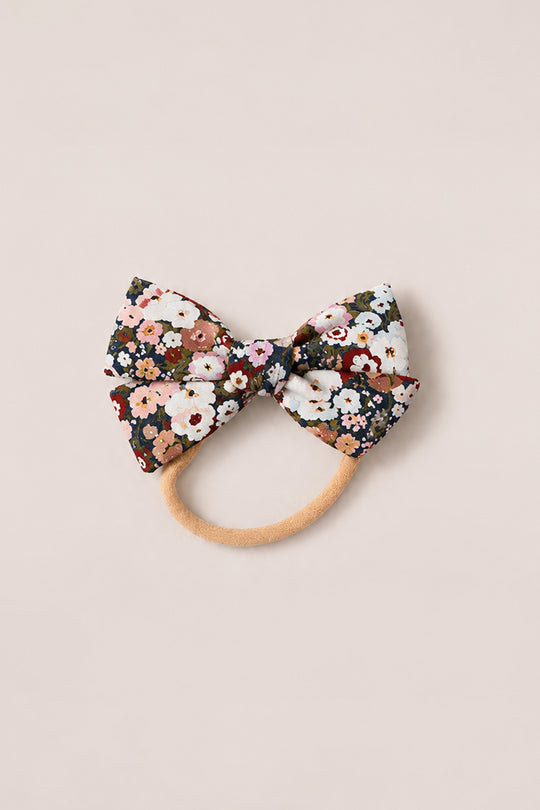 Introducing the Hair Bows in Midnight Garden by Ivy City Co—an elegant hair accessory featuring a large fabric bow adorned with a vibrant floral pattern in red, pink, white, and green. Reminiscent of Ivy City Co’s signature femininity, this charming hair tie is set on a beige elastic band and elevates any look with the enchanting spirit of Midnight Garden. Photographed delicately on a light background.