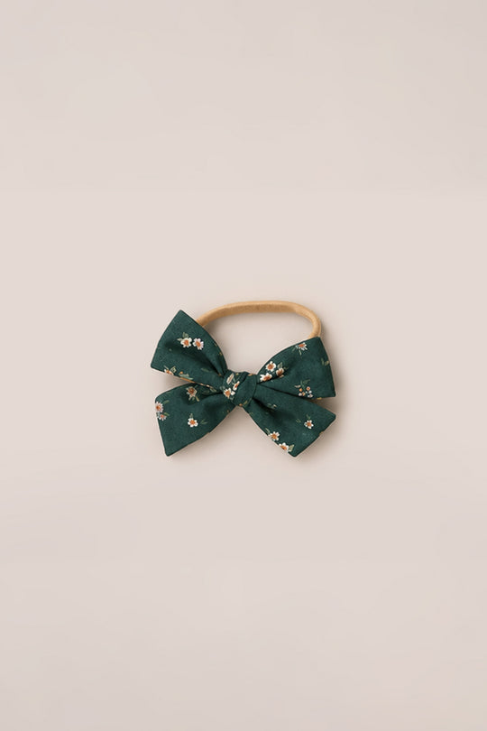 The Ivy City Co Hair Bows in Evergreen Lovelorn Ditsy feature a green fabric bow with a delicate floral pattern, attached to a beige elastic headband and displayed on a simple light background—a charming accessory inspired by Ivy City style.