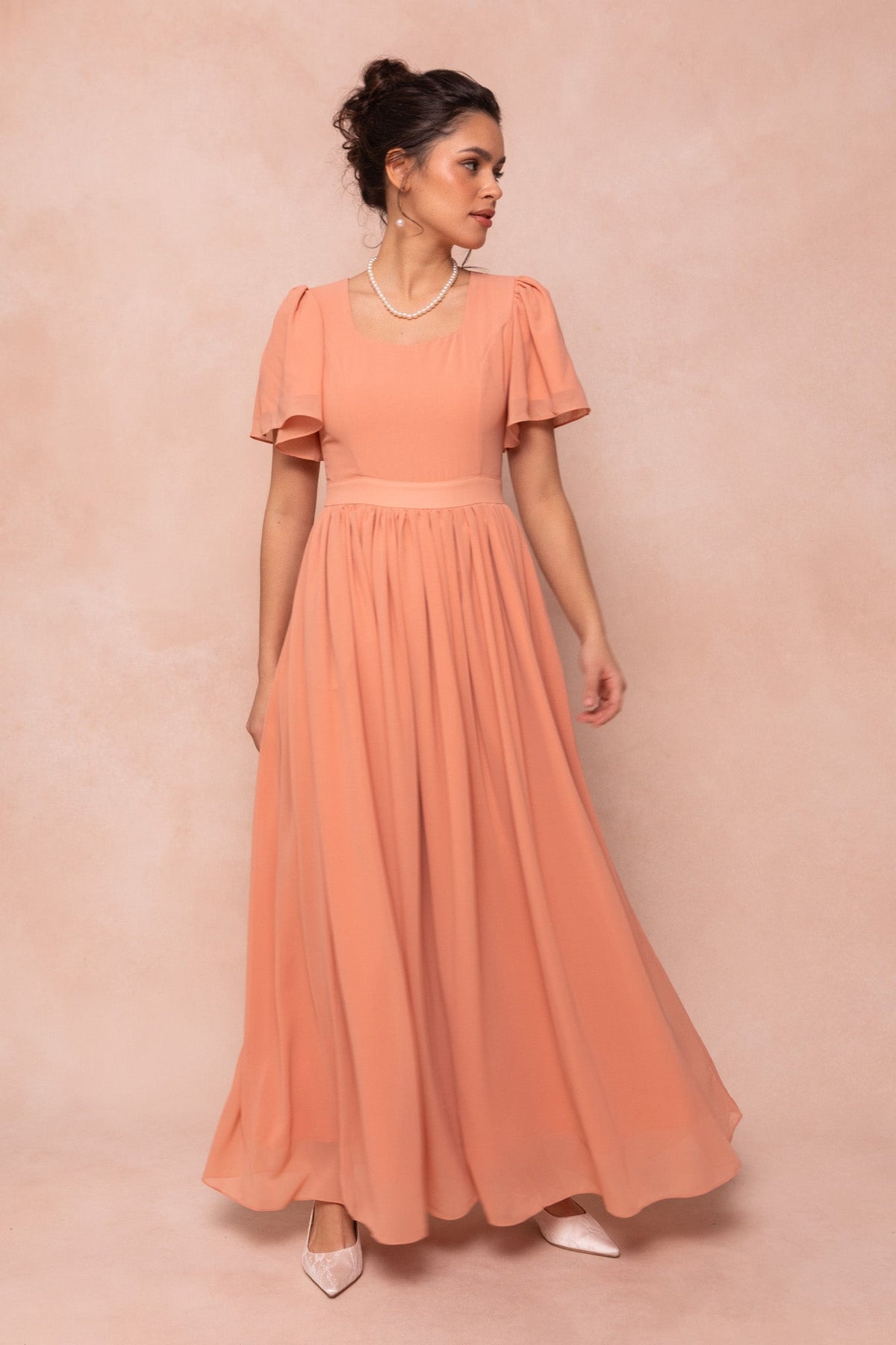 A woman wearing the Ivy City Co Abbie Dress in Apricot Crush, a short-sleeved maxi dress, stands against a soft beige background looking to her left. She accessorizes with pearl earrings, a necklace, and pointed-toe shoes.