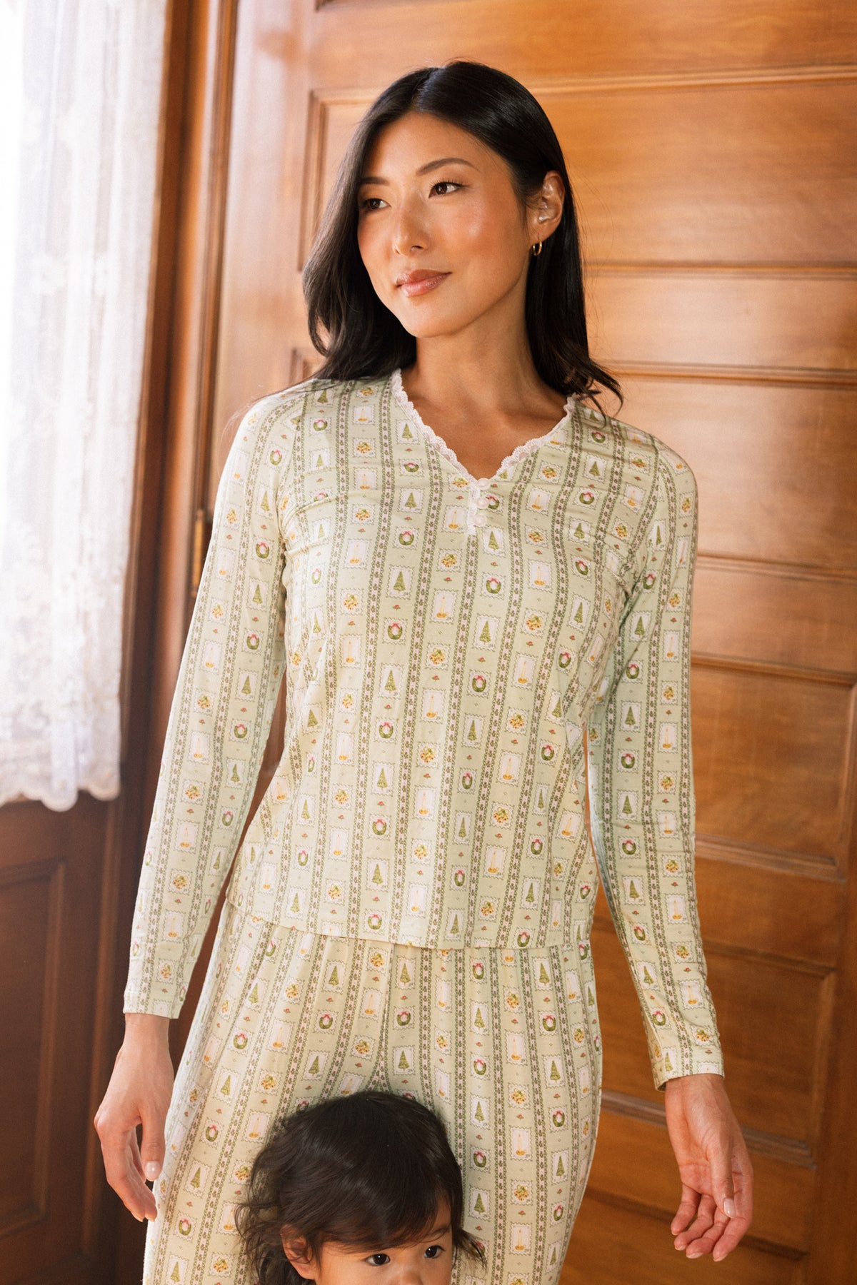 A woman wears the So Soft Henley Pajama Set in Merry Mail Linear by Ivy City Co, standing indoors by a wooden door as light streams through lace curtains; a child is partially visible, holding her leg.