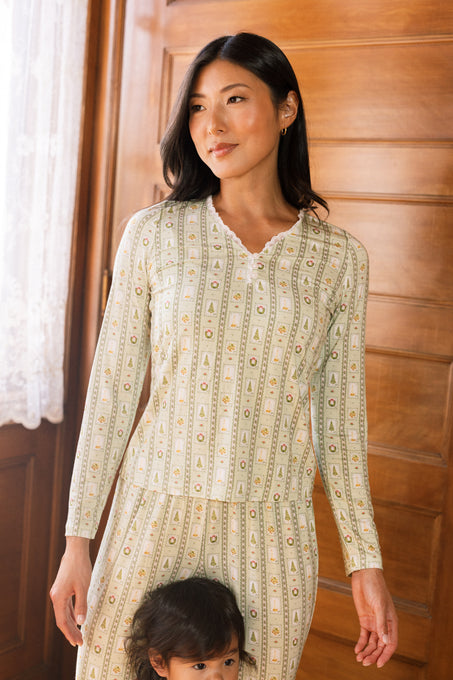 A woman wears the So Soft Henley Pajama Set in Merry Mail Linear by Ivy City Co, standing indoors by a wooden door as light streams through lace curtains; a child is partially visible, holding her leg.
