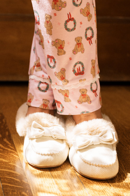 A person wears the So Soft Henley Pajama Set in Teddy Bear Wreaths by Ivy City Co—pink pajamas with teddy bear and wreath prints—standing in white fuzzy slippers with bows on a wooden floor.