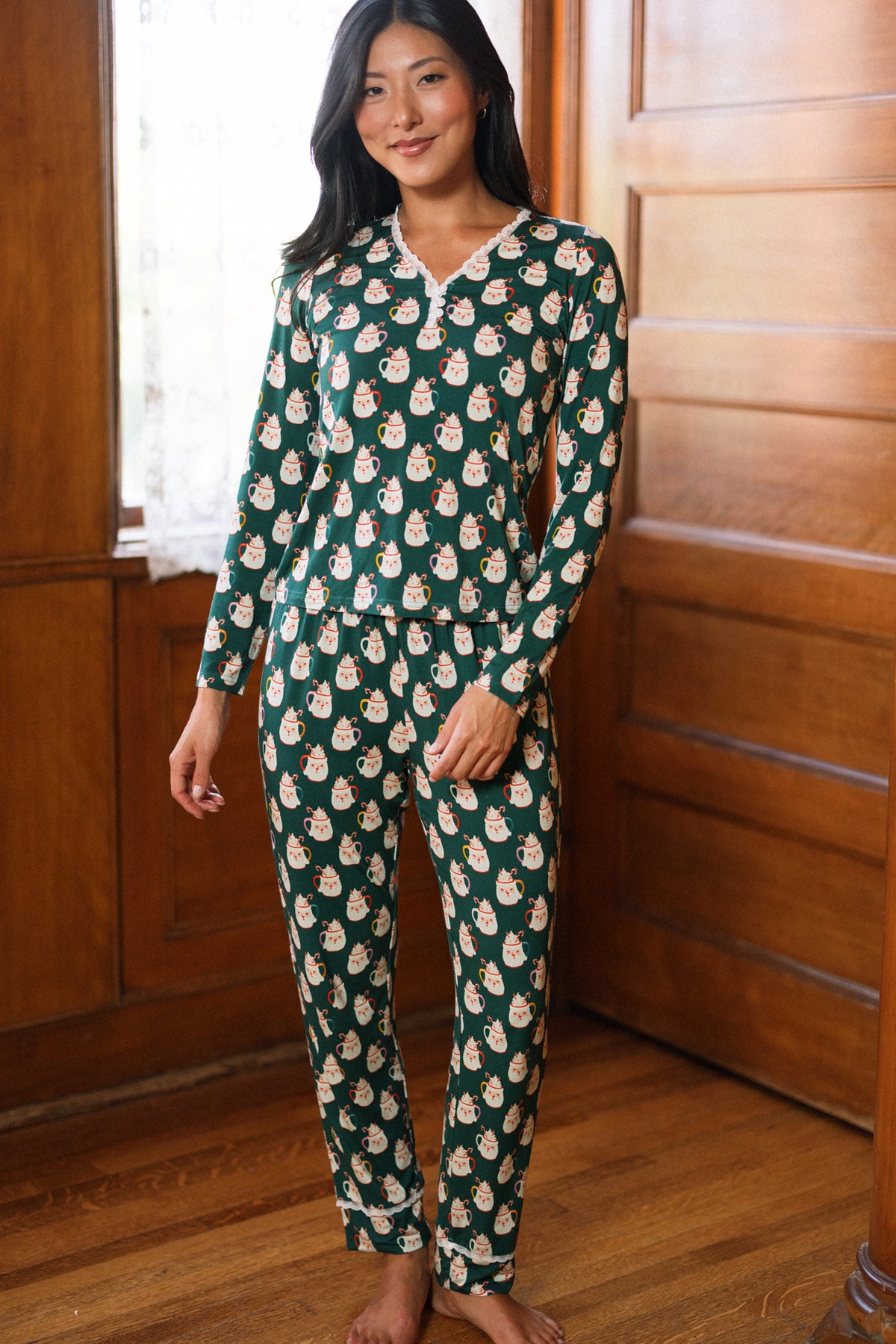 A woman stands barefoot on a wooden floor, wearing the So Soft Henley Pajama Set in Very Merry Santa by Ivy City Co, in a room with wood-paneled walls and white lace curtains.