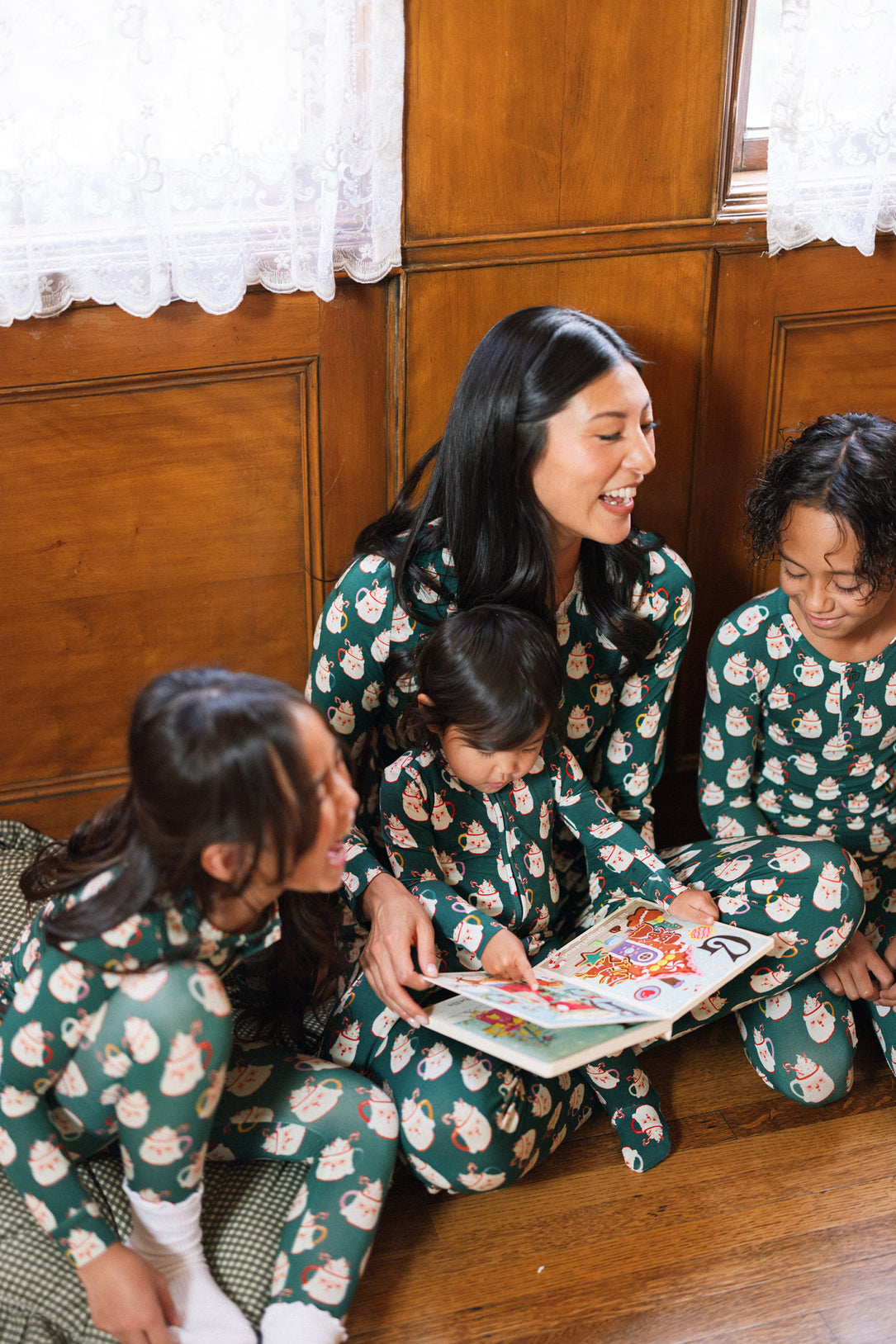 A woman and three children wear the So Soft Henley Pajama Set in Very Merry Santa by Ivy City Co as they sit on a wooden floor, smiling and reading a picture book together by a window with lace curtains.