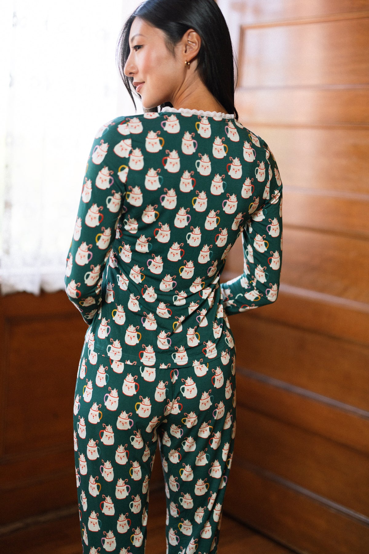 A woman stands in a wooden room, wearing the So Soft Henley Pajama Set in Very Merry Santa by Ivy City Co, looking over her shoulder as natural light shines through the window.