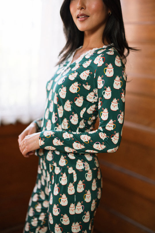 A woman stands indoors wearing the So Soft Henley Pajama Set in Very Merry Santa by Ivy City Co, partially turned to the side and smiling softly with natural light streaming through a window behind her.