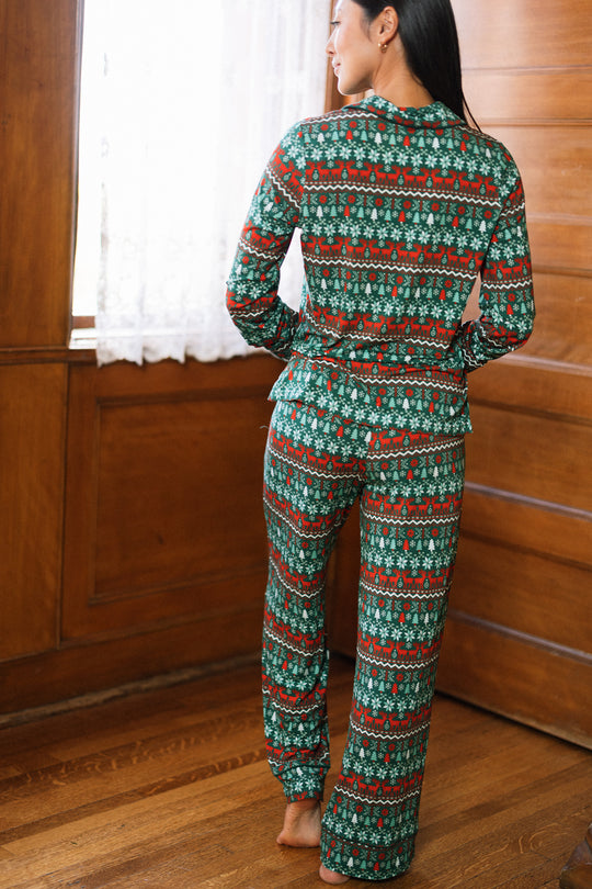 A person stands barefoot indoors, wearing the So Soft Pajama Set in Alpine Noel by Ivy City Co, featuring green with red and white festive patterns. The room has wood floors, paneled walls, and a window with lace curtains.