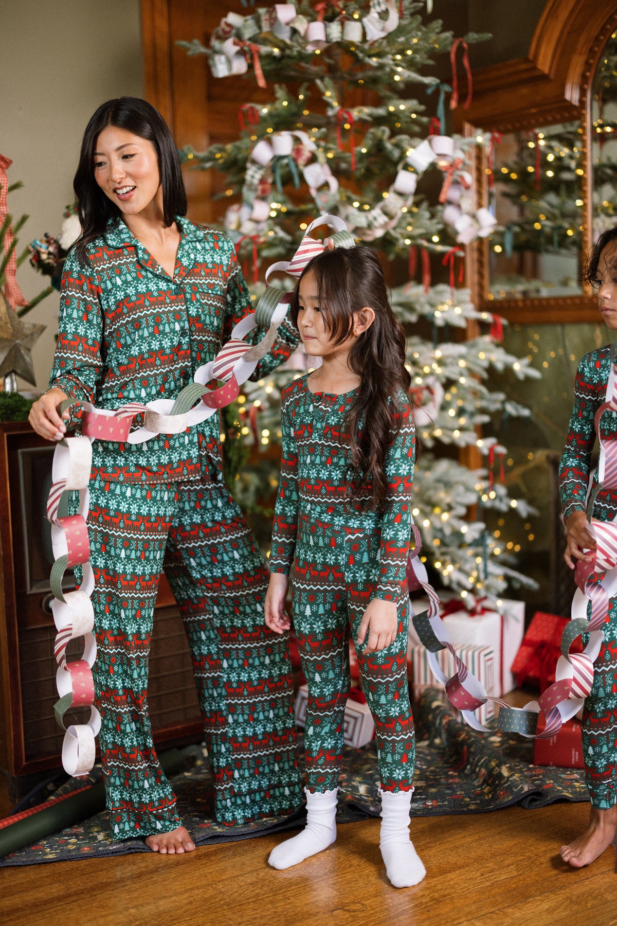 A woman and two children in matching Ivy City Co So Soft Pajama Set in Alpine Noel decorate a Christmas tree with paper chains, surrounded by wrapped gifts and glowing festive lights.