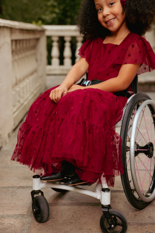 Young girl in a red dress with black polka dots sitting in a wheelchair outdoors.