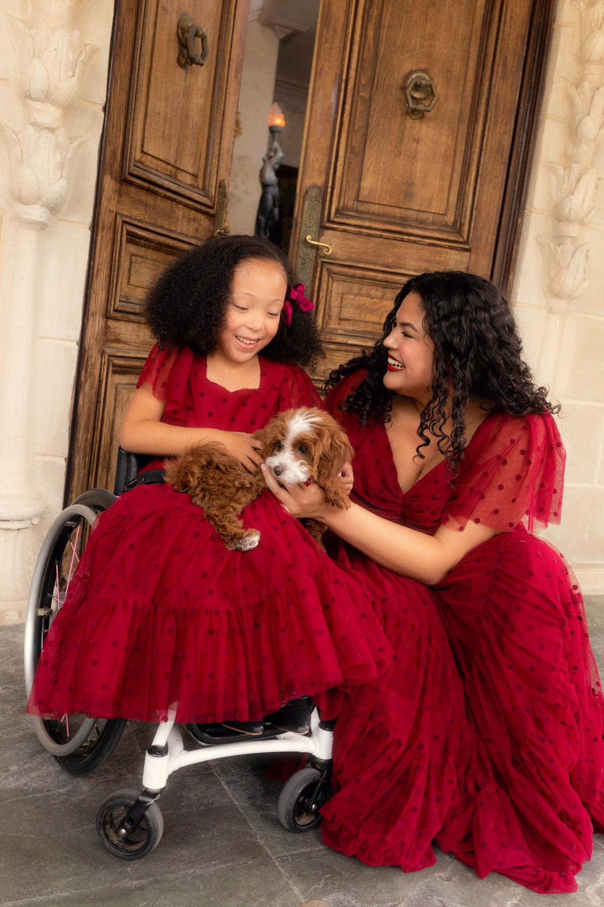 Two women in red dresses with a dog in front of a wooden door.