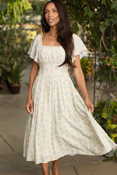 A woman wears the Ivy City Co Sonoma Dress in Buttercream Porcelain Fleur, a light yellow floral midi with short sleeves, outdoors amid greenery—smiling softly and gently holding her dress, she’s the picture of a summer wedding guest.