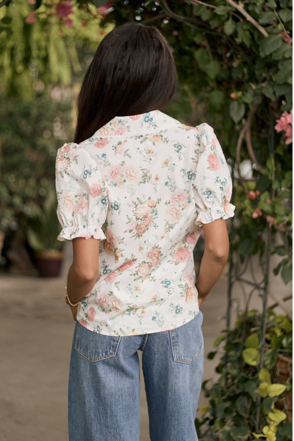 A woman with long dark hair stands outdoors, wearing the Ivy City Co Sophia Blouse in Countryside Peony Blossom and blue jeans. She faces away, surrounded by greenery and pink flowers.