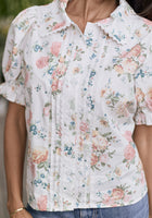 Someone wearing the Ivy City Co Sophia Blouse in Countryside Peony Blossom, an ivory floral blouse with lace trim, pleats, front buttons, puffed short sleeves, paired with blue jeans.