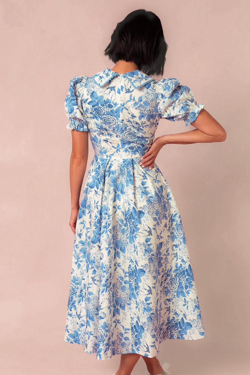 A woman with short dark hair stands with her back to the camera, modeling Ivy City Co's Sophia Dress in Blue Floral—a nursing-friendly midi dress featuring puffed sleeves and a fitted waist—set against a neutral background.