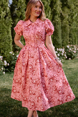 A woman stands outdoors on grass, holding the skirt of the Sophia Dress in Desert Heritage Rose by Ivy City Co—a pink cotton floral dress with red rose patterns and puffed short sleeves. Green trees and blooming flowers are in the background.
