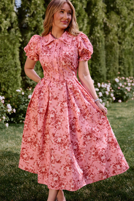 A woman stands outdoors on grass, holding the skirt of the Sophia Dress in Desert Heritage Rose by Ivy City Co—a pink cotton floral dress with red rose patterns and puffed short sleeves. Green trees and blooming flowers are in the background.