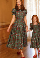 A woman and a young girl hold hands and walk indoors on a wooden floor, both wearing matching Ivy City Co “Sophia Dress in Evergreen Merry Ditsy” dresses with puff sleeves and red shoes.