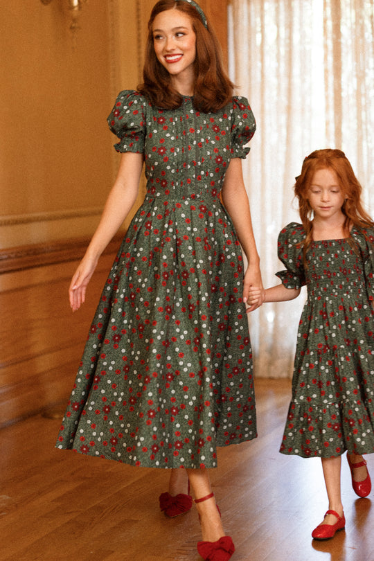 A woman and a young girl hold hands and walk indoors on a wooden floor, both wearing matching Ivy City Co “Sophia Dress in Evergreen Merry Ditsy” dresses with puff sleeves and red shoes.