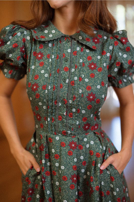 A person wearing the Ivy City Co Sophia Dress in Evergreen Merry Ditsy—a vintage-inspired dress with a green base and red and white floral pattern, short puff sleeves, a wide collar, and hands tucked in the dress’s pockets. The softly blurred background accentuates the look.