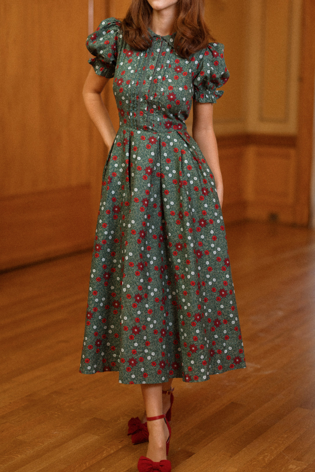 A woman stands indoors on a wooden floor wearing the Ivy City Co Sophia Dress in Evergreen Merry Ditsy—a green, floral-patterned, knee-length vintage dress with short puff sleeves and pockets. She pairs it with red shoes featuring large bows. Her face is not visible.