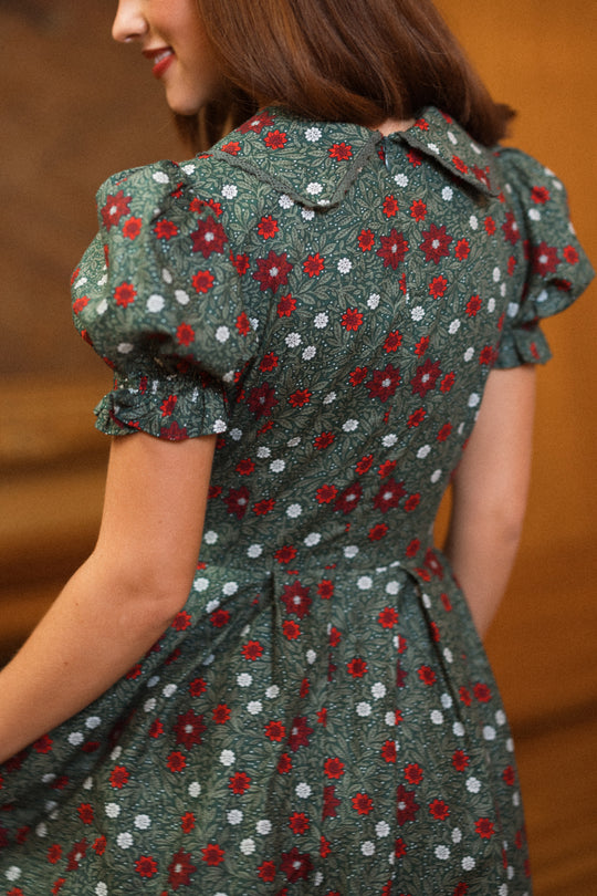 A woman in the Ivy City Co Sophia Dress in Evergreen Merry Ditsy stands with her back to the camera, showcasing its green fabric adorned with red and white florals, puff sleeves, and fitted waist.