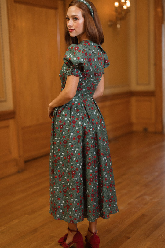 A woman wearing the Ivy City Co Sophia Dress in Evergreen Merry Ditsy, paired with red shoes and a coordinating headband, stands on a wooden floor in an elegant, warmly lit room.