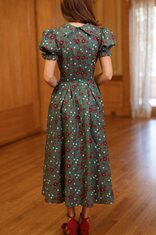 A woman stands indoors on a wooden floor with her hair down, facing away from the camera, wearing the Ivy City Co Sophia Dress in Evergreen Merry Ditsy—an ankle-length vintage-inspired green dress featuring red and white floral patterns and puffed sleeves.