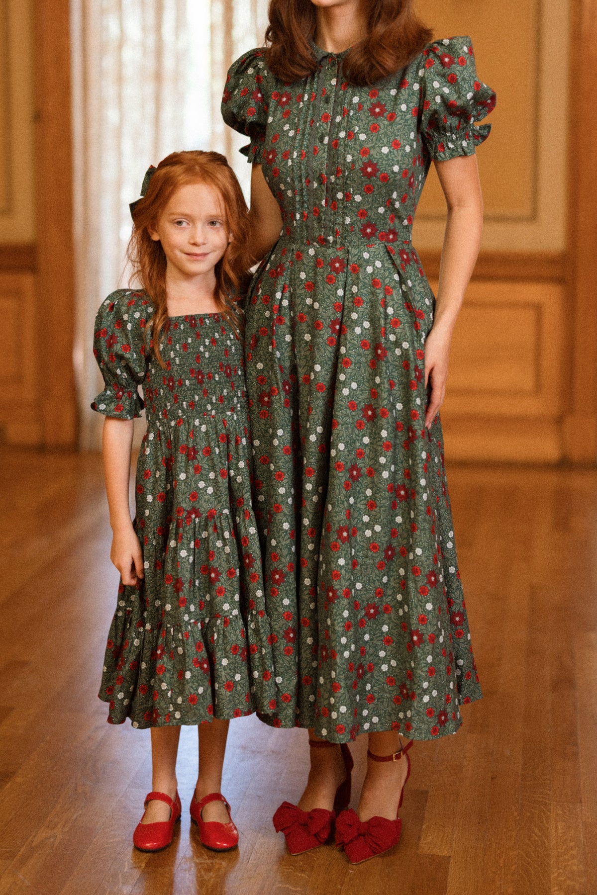 A young girl with long red hair and an adult woman stand together indoors, both wearing the charming Ivy City Co “Sophia Dress in Evergreen Merry Ditsy” with puff sleeves and a festive red and white floral pattern, paired with matching red shoes. The scene is set in a cozy room with wooden floors and paneling.