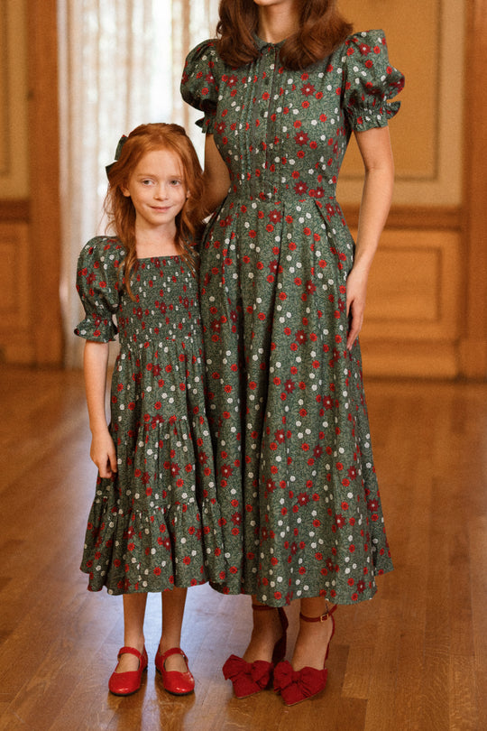 A young girl with long red hair and an adult woman stand together indoors, both wearing the charming Ivy City Co “Sophia Dress in Evergreen Merry Ditsy” with puff sleeves and a festive red and white floral pattern, paired with matching red shoes. The scene is set in a cozy room with wooden floors and paneling.