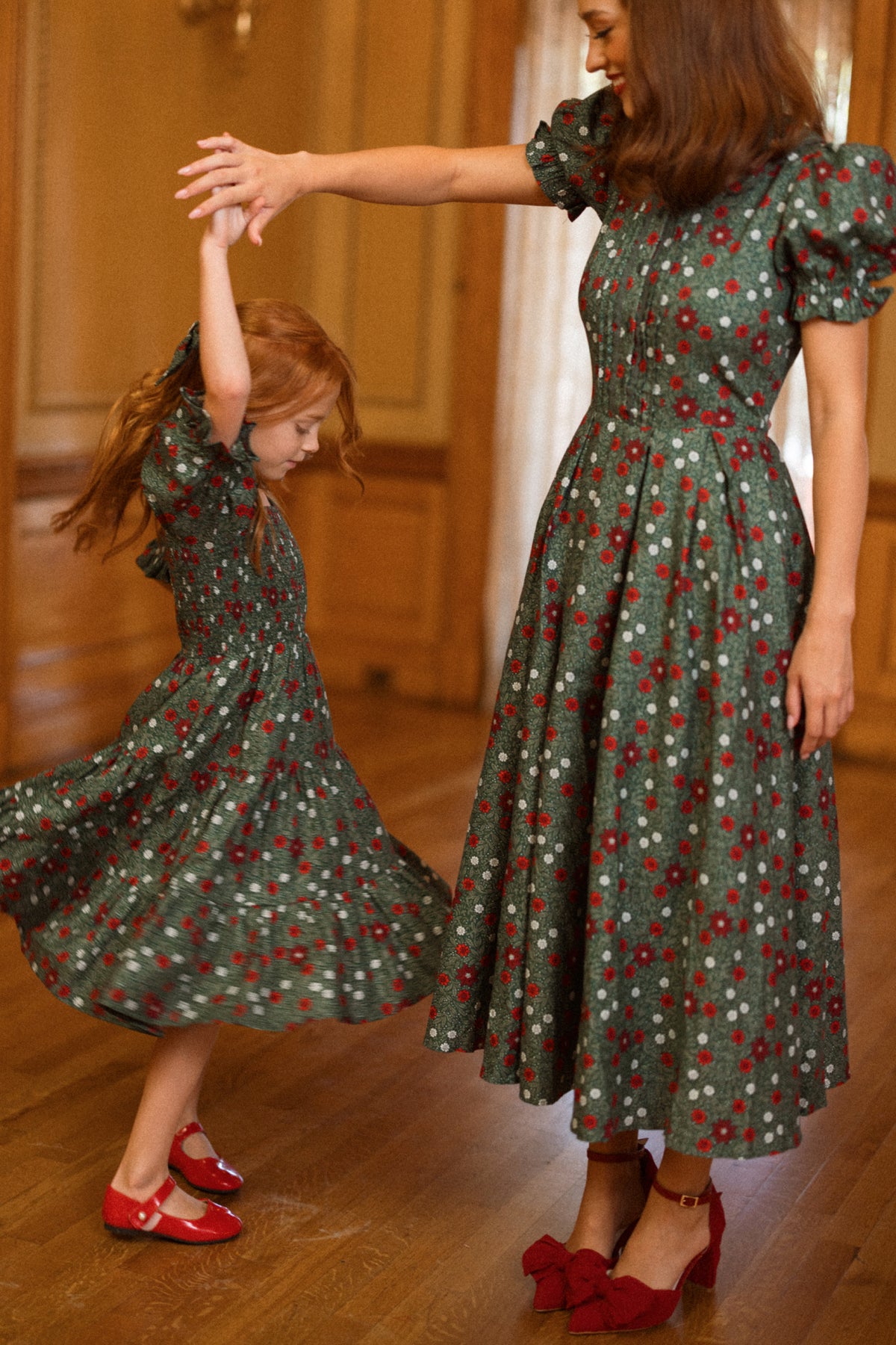 A woman and a young girl dressed in matching Ivy City Co Sophia Dresses in Evergreen Merry Ditsy dance joyfully indoors, smiling as the girl twirls and her red hair and dress spin. Both wear red shoes that complement their coordinated holiday outfits, adding a charming touch to the festive scene.