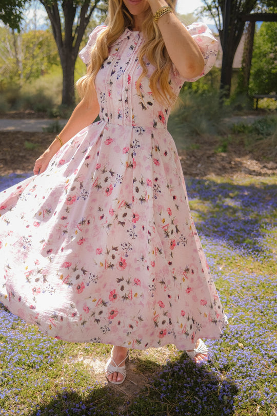 A person with long wavy hair, wearing the Ivy City Co Sophia Dress in Roselyn Pink and white sandals, twirls outdoors on a field of purple flowers, holding the hem. Their face is not visible.