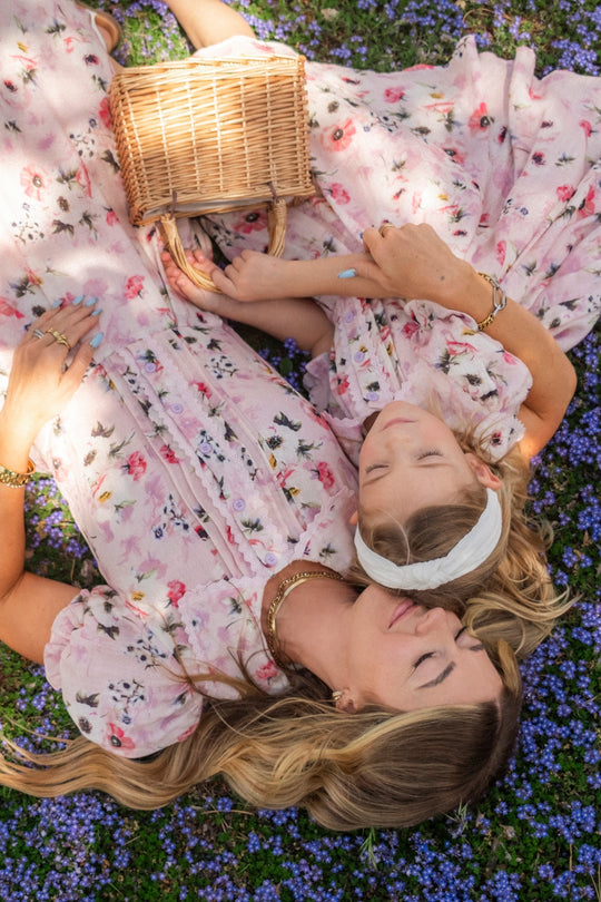 Two people in matching Ivy City Co Sophia Dress in Roselyn Pink lie on grass dotted with small purple flowers, holding a wicker basket. They relax close together, enjoying a peaceful outdoor moment.