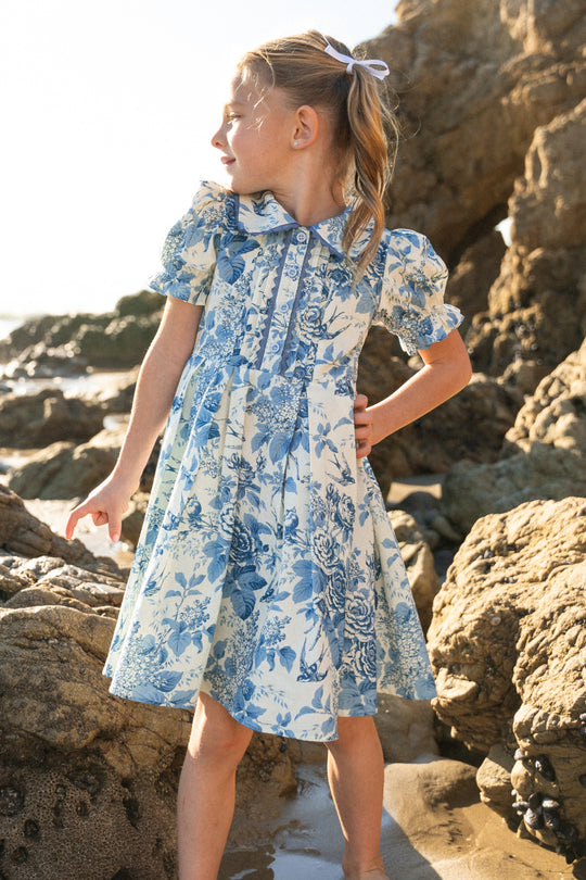 A young girl wears the Ivy City Co Mini Sophia Dress in Blue Floral, featuring puffed sleeves, as she stands on rocky terrain. With a ribbon in her sunlit hair, she poses with one hand on her hip and the other pointing to the side.