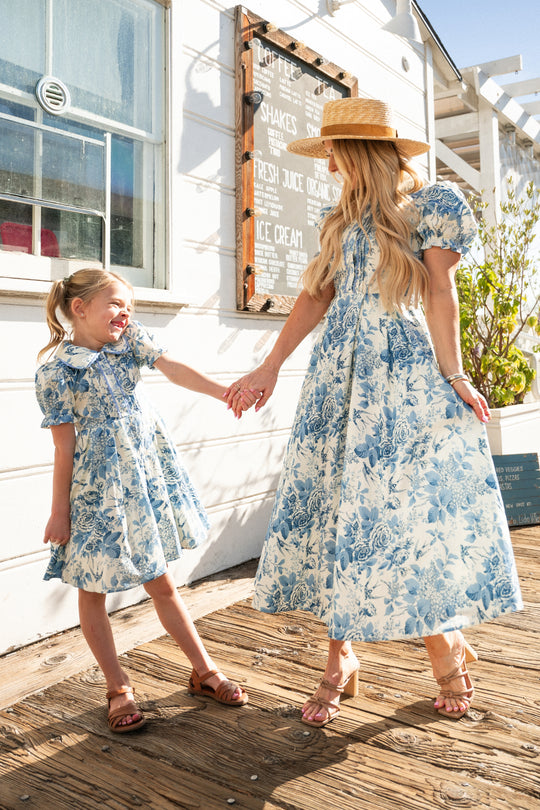 A woman and young girl, both in Ivy City Co's Mini Sophia Dress in Blue Floral, hold hands and smile outside a white building on a sunny day, with the woman adding flair with a straw hat as they enjoy time together.