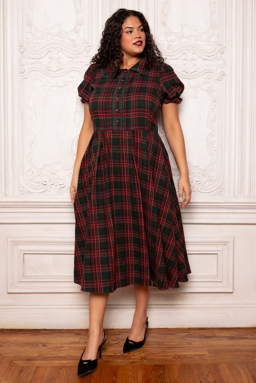 Woman wearing a red and green plaid dress against a white wall.