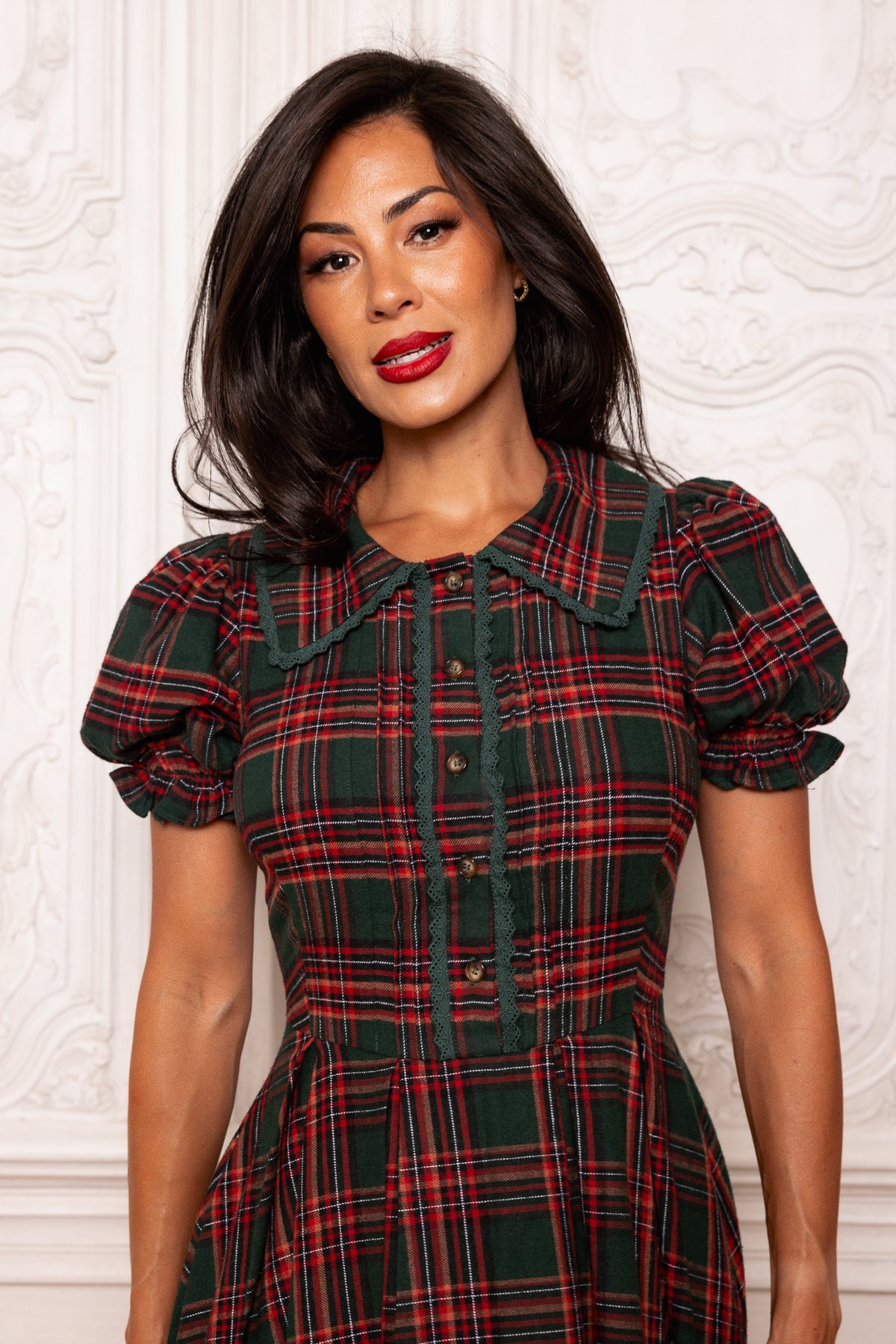 Woman wearing a red and green plaid dress against a white wall.