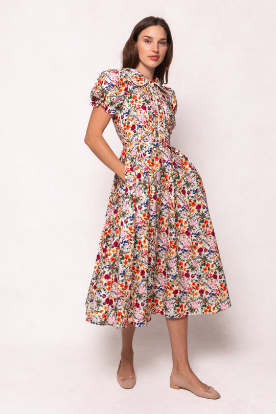 A woman models the Ivy City Co Sophia Dress in Multicolor Floral, a cotton midi dress with puffed short sleeves, pockets, a fitted waist, and vibrant floral print—styled with beige flats against a light background.
