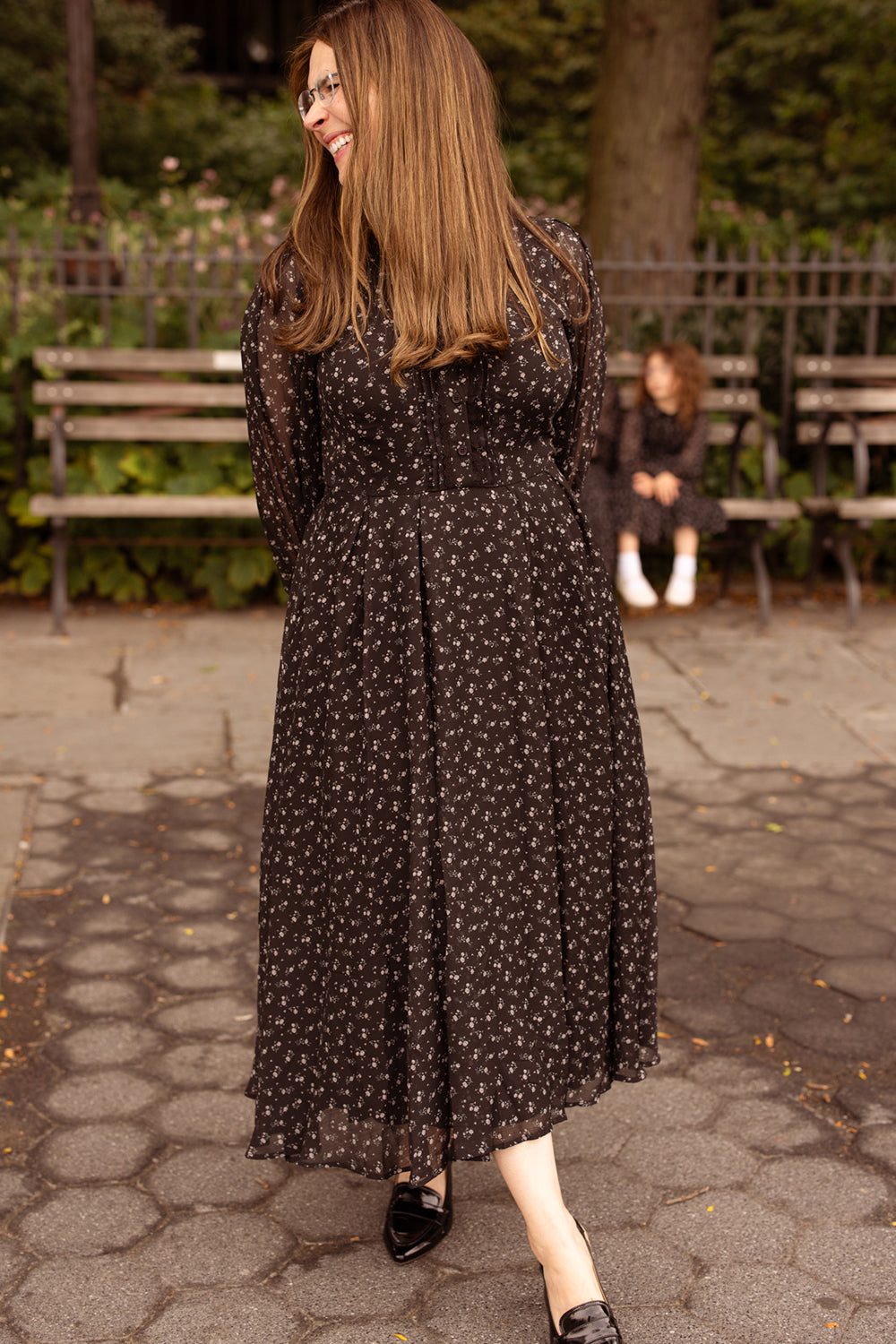A woman in the Ivy City Co Sophia Long Sleeve Dress in Noir Ditsy Chiffon smiles and looks to the side in a park. In the background, a child sits on a bench, also dressed in black. This nursing-friendly, tea-length dress blends elegance and comfort, making it perfect for any outdoor outing.