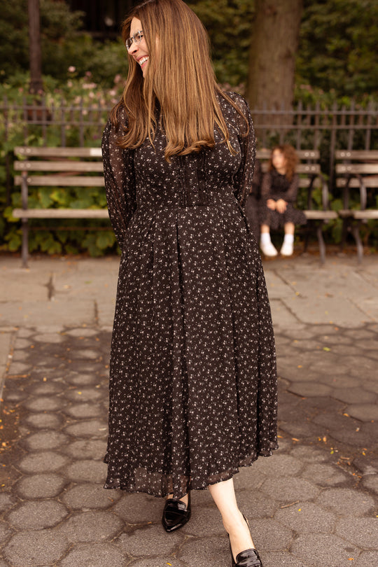 A woman in the Ivy City Co Sophia Long Sleeve Dress in Noir Ditsy Chiffon smiles and looks to the side in a park. In the background, a child sits on a bench, also dressed in black. This nursing-friendly, tea-length dress blends elegance and comfort, making it perfect for any outdoor outing.