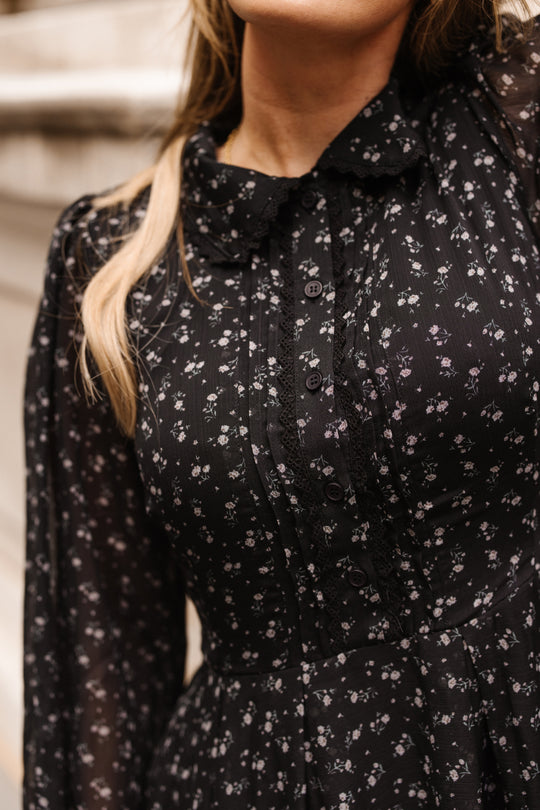 A woman stands outdoors wearing the Sophia Long Sleeve Dress in Noir Ditsy Chiffon by Ivy City Co, a black tea-length dress featuring a floral pattern and lace accents. The photo is cropped to show her upper body and part of her face.
