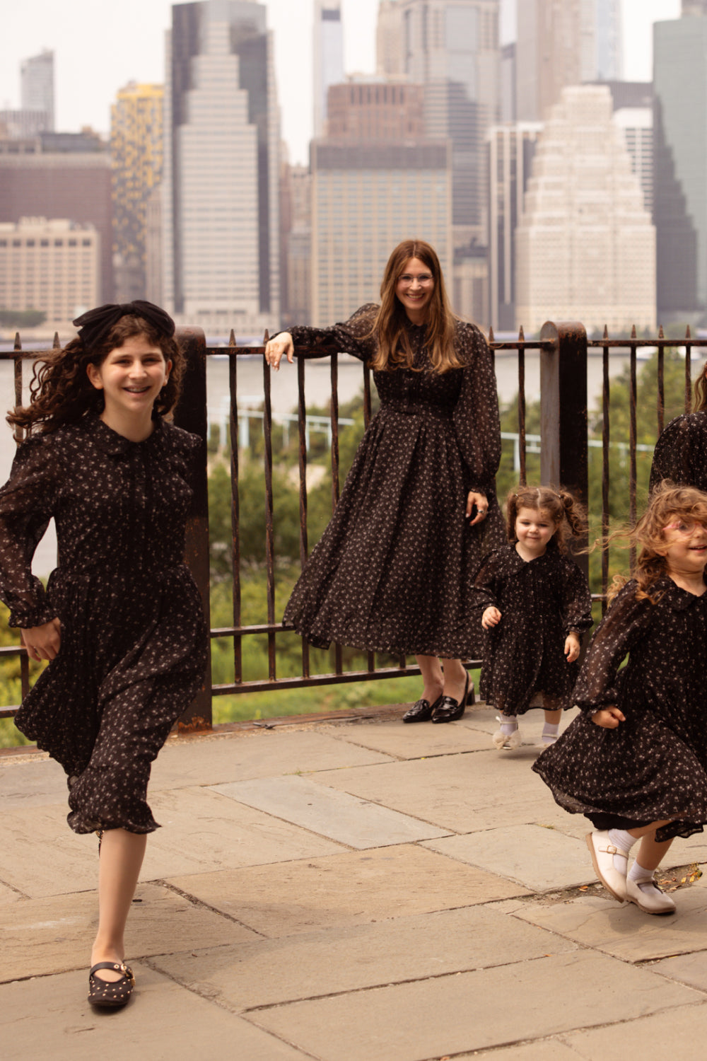 Four girls and a woman, all dressed in matching Sophia Long Sleeve Dress in Noir Ditsy Chiffon by Ivy City Co, stand and play on a paved area with a city skyline and green trees in the background.