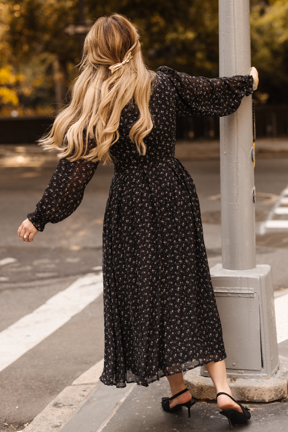 A woman with long blonde hair styled in a ribbon wears the Ivy City Co Sophia Long Sleeve Dress in Noir Ditsy Chiffon and heels, holding onto a street pole as she walks. Trees and a crosswalk appear in the background.