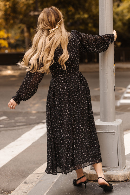 A woman with long blonde hair styled in a ribbon wears the Ivy City Co Sophia Long Sleeve Dress in Noir Ditsy Chiffon and heels, holding onto a street pole as she walks. Trees and a crosswalk appear in the background.