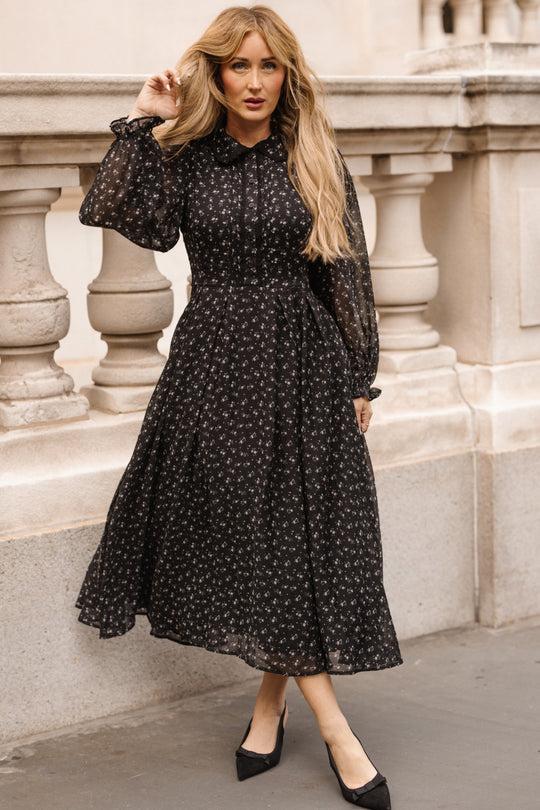 A woman with long blonde hair wears the Ivy City Co Sophia Long Sleeve Dress in Noir Ditsy Chiffon—a nursing-friendly, black floral tea-length dress—paired with black heels, standing outdoors near a stone railing with ornate columns.
