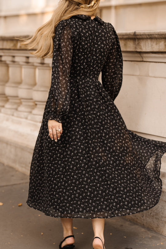 A woman with long blonde hair wears the Ivy City Co Sophia Long Sleeve Dress in Noir Ditsy Chiffon, a nursing-friendly black floral tea-length dress, paired with black sandals as she walks outdoors near a stone railing.