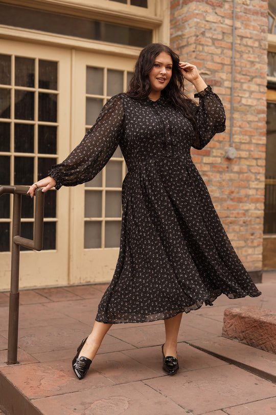 A woman wearing the Ivy City Co Sophia Long Sleeve Dress in Noir Ditsy Chiffon poses outdoors on a stone walkway, holding a handrail and touching her hair, with a brick wall and windows in the background. This nursing-friendly, tea-length dress blends elegance with practical comfort.