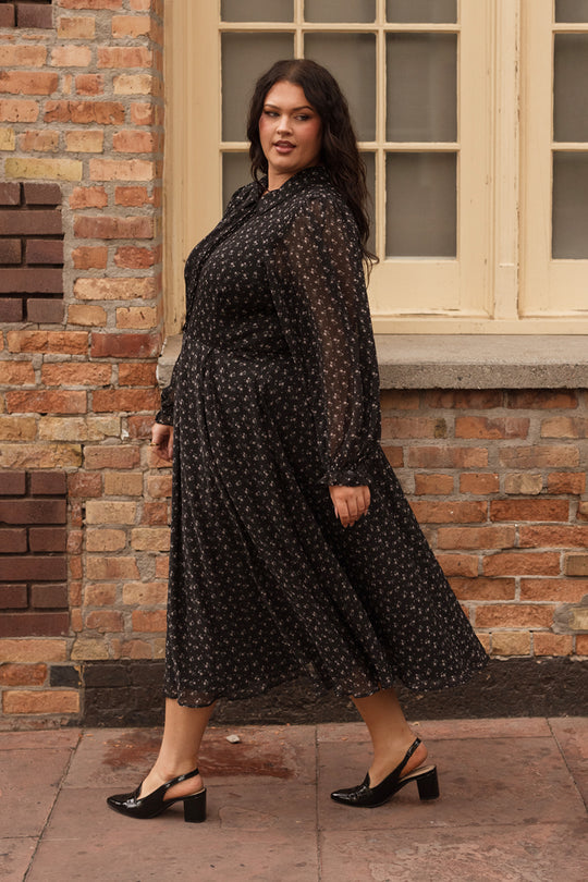 A woman wearing the Ivy City Co Sophia Long Sleeve Dress in Noir Ditsy Chiffon, a nursing-friendly tea-length dress with a black ditsy pattern, pairs it with black heels as she walks past a brick wall and large cream window, looking confidently at the camera.