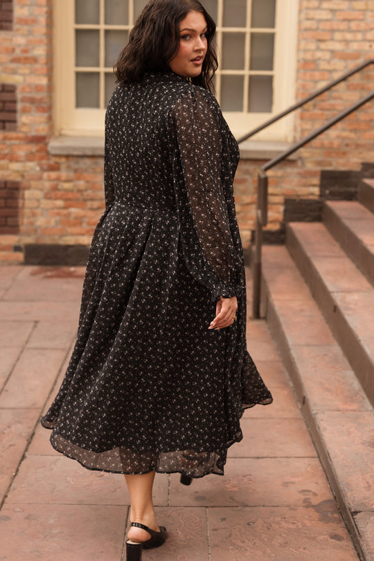 A woman in the Ivy City Co Sophia Long Sleeve Dress in Noir Ditsy Chiffon looks over her shoulder while walking outdoors on a stone path near brick steps and a window.